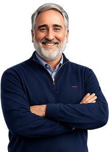 Smiling Ed Bejarana with short grey hair and a well-groomed grey beard, wearing a navy blue quarter-zip sweater over a light blue button-down shirt, standing with arms crossed against a white background.
